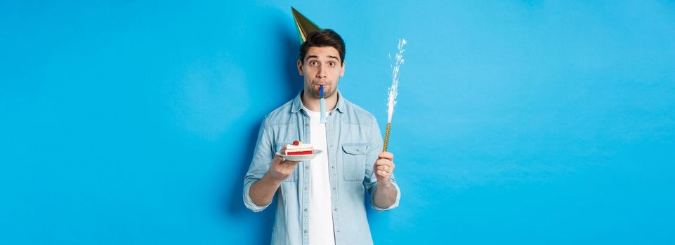 Funny guy celebrating birthday, holding b-day cake, firework and wearing party hat, standing over blue background.