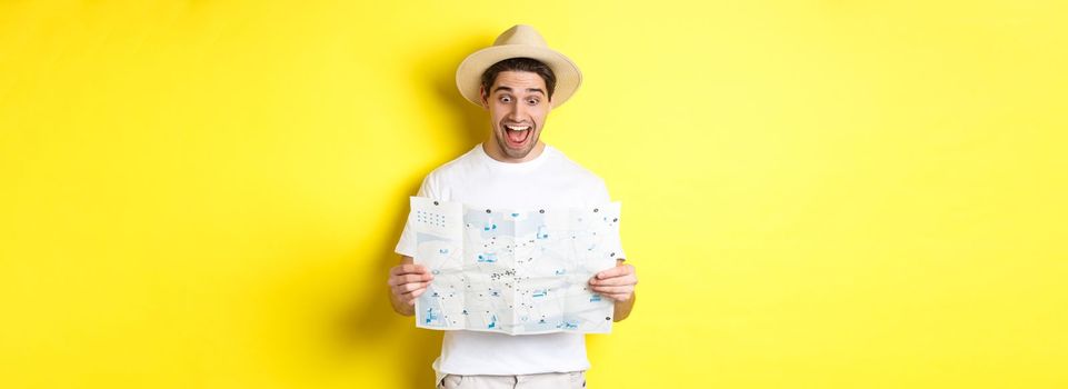 Travelling, vacation and tourism concept. Man tourist looking happy at map with sighsteeings, explore city, standing over yellow background.