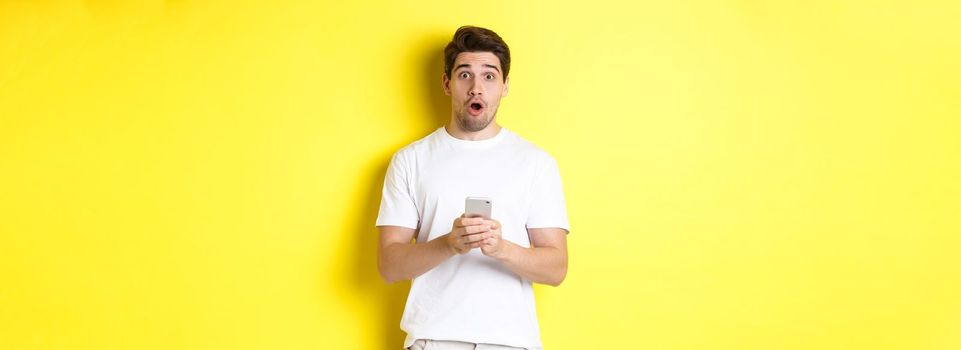Man looking surprised, using smartphone, open mouth and saying wow, standing against yellow background. Copy space