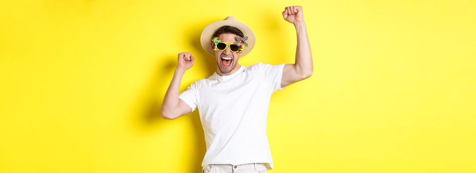 Concept of tourism and lifestyle. Happy lucky guy winning trip, rejoicing and wearing holiday outfit, summer hat and sunglasses, yellow background.