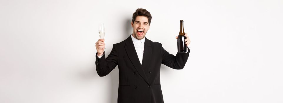 Concept of holidays, party and celebration. Handsome man in trendy suit having fun, holding bottle and glass of champagne, standing over white background.