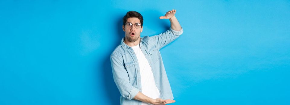 Confused handsome man showing big size, shaping large box and looking amazed, standing over blue background.