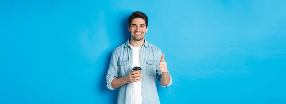 Handsome young man showing thumb-up and drinking coffee, recommending cafe takeaway, standing over blue background.