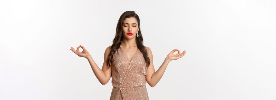 Christmas party and celebration concept. Calm and patient young woman in stylish dress meditating, feeling zen, standing over white background.