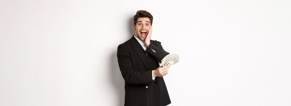 Image of rich and happy man in black suit, winning prize, holding money and looking excited at camera, standing over white background.