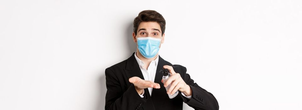 Concept of covid-19, business and social distancing. Image of handsome businessman in trendy suit and medical mask, cleaning hands with hand sanitizer, standing over white background.