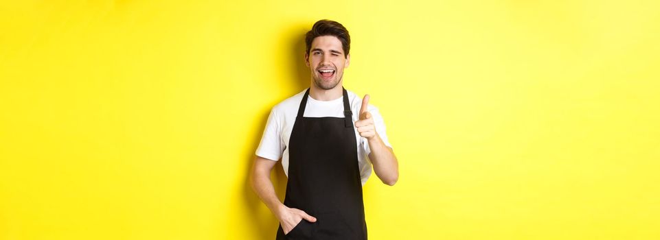 Handsome barista in black apron welcome guests to coffee shop, pointing finger gun and winking, standing against yellow background.