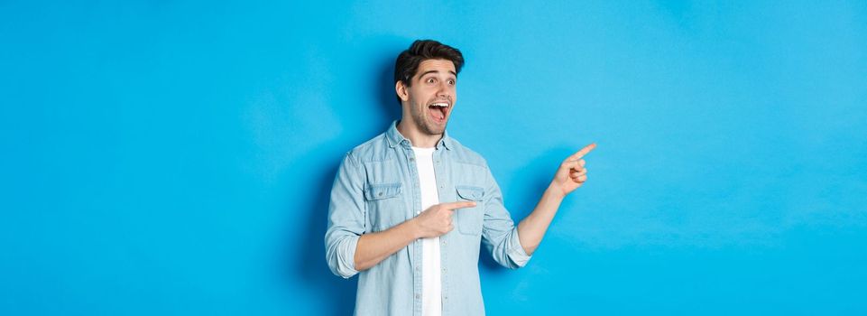 Portrait of handsome man looking and pointing fingers left with excitement, standing against blue background.