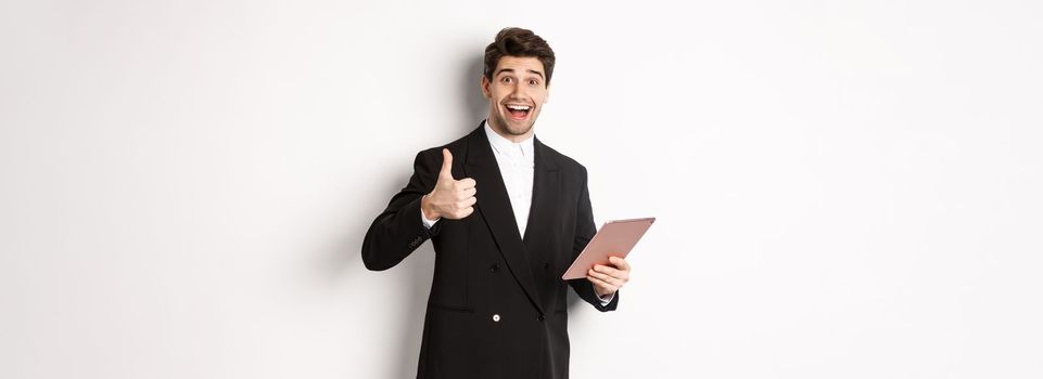 Portrait of handsome businessman in suit, showing thumb-up and making a compliment, praising something on digital tablet, standing over white background.