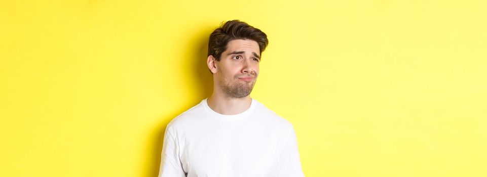 Reluctant guy in white t-shirt looking left, grimacing skeptical and displeased, standing over yellow background.