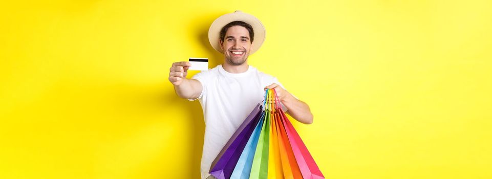 Smiling young man buying things with credit card, holding shopping bags and looking happy, standing over yellow background.