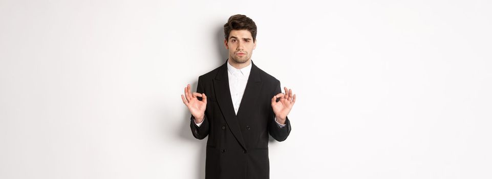 Concept of new year party, celebration and lifestyle. Portrait of confident good-looking man in black suit, showing okay sign and approve something, standing over white background.