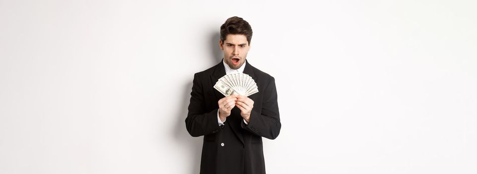 Portrait of handsome businessman in trendy suit, looking startled at money, won prize, standing over white background.