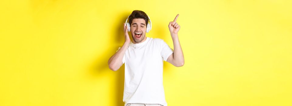 Happy man listening music in headphones, pointing finger at promo offer for black friday, standing over yellow background.