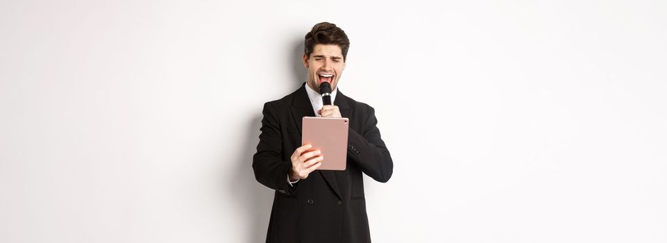 Image of handsome man in black suit, singing karaoke on digital tablet, holding microphone, standing over white background.