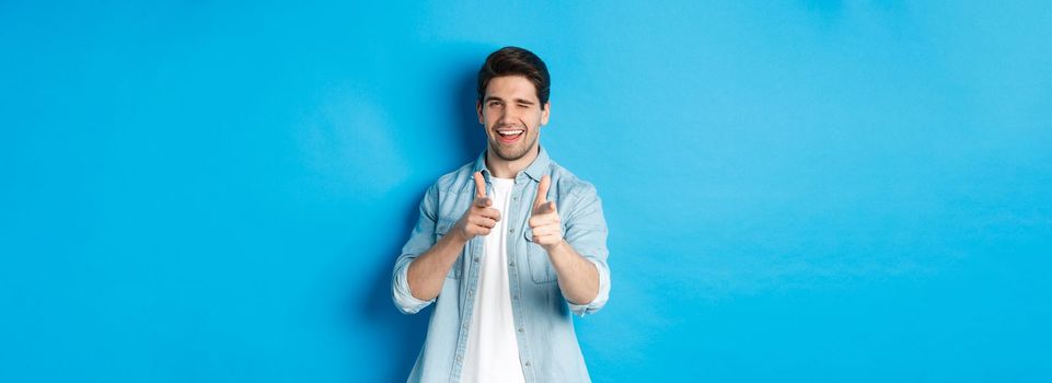 Cheeky handsome guy pointing fingers at you, winking flirty, standing in casual outfit against blue background.