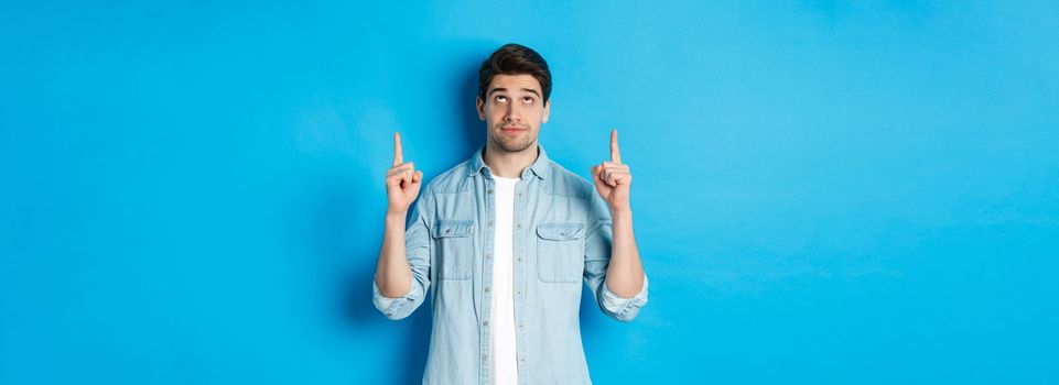 Portrait of displeased and skeptical male model pointing fingers up, looking at something unpleasant, standing against blue background.