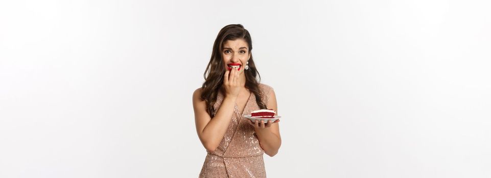 Image of young woman hesitating to eat cake on party, biting fingernails from temptation, standing over white background in elegant dress.