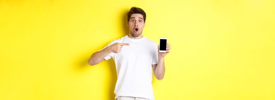 Impressed guy check out smartphone screen, pointing finger at display and gasping amazed, standing over yellow background. Copy space