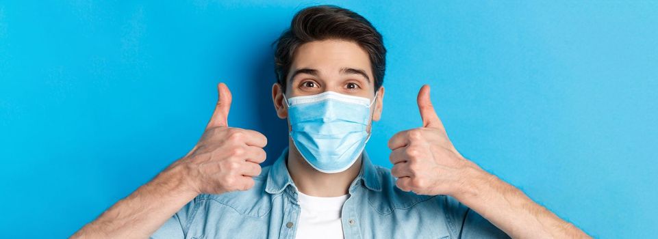 Concept of covid-19, pandemic and quarantine. Close-up of cheerful young man in medical mask smiling, showing thumbs up in approval, like and agree, standing over blue background.