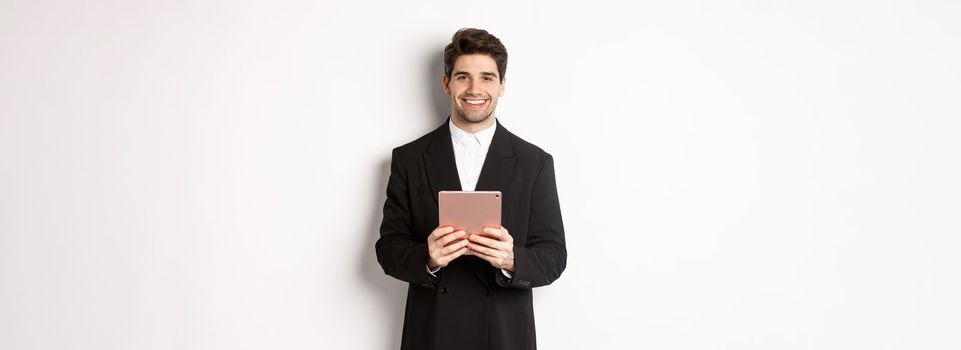 Image of handsome businessman in trendy suit, holding digital tablet and smiling, standing against white background.