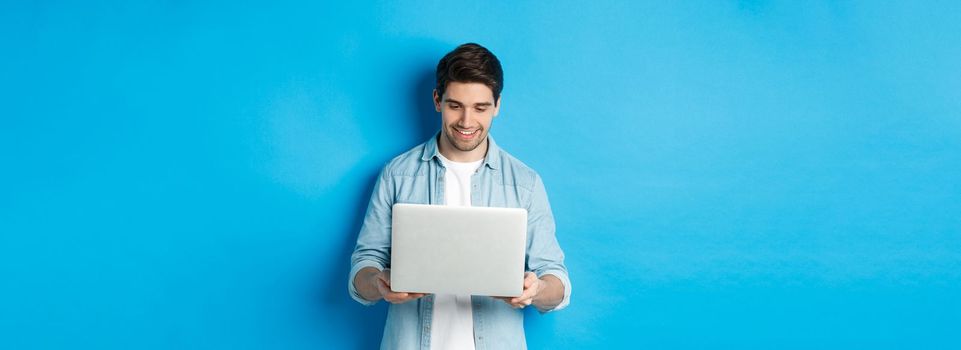 Handsome man working on laptop, smiling and looking at screen satisfied, standing against blue background.