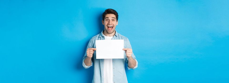 Excited man making fantastic announcement, showing logo or sign on blank piece of paper, standing over blue background.