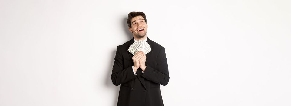 Image of handsome dreamy man in black suit, holding money and looking at upper left corner, thinking about shopping, standing over white background.