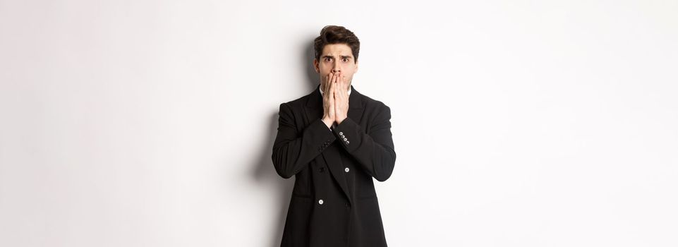 Portrait of shocked handsome businessman in suit, reacting to terrible situation, gasping and covering mouth with hands, standing startled against white background.