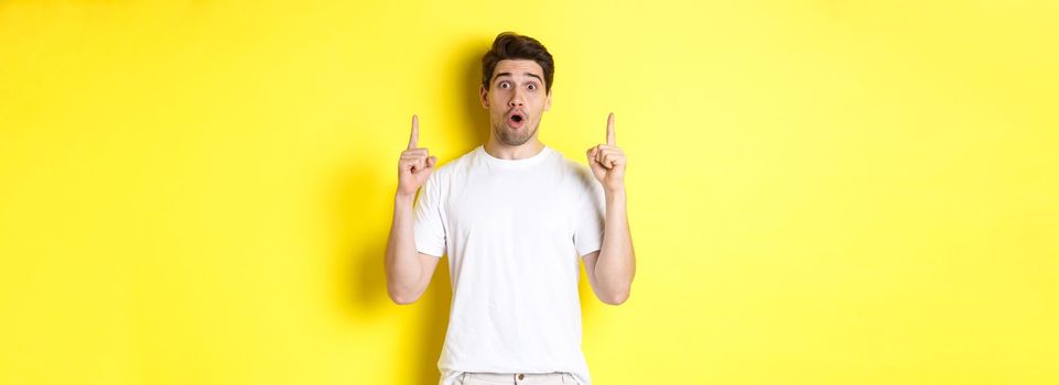 Surprised handsome guy in white t-shirt, pointing fingers up, interested about advertisement, standing against yellow background.