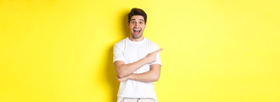 Happy man pointing finger left, showing advertisement on copy space, smiling amused, standing in white clothes against yellow background.