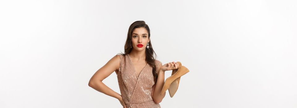 Party and celebration concept. Sassy and beautiful woman in luxury dress and red lipstick, holding pair of heels and looking unbothered, standing over white background.
