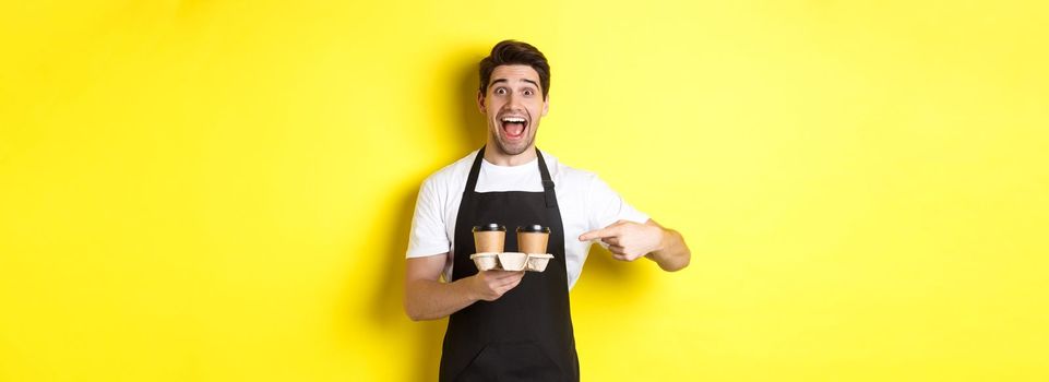 Excited barista in black apron pointing fingers at takeaway coffee cups, standing against yellow background happy.