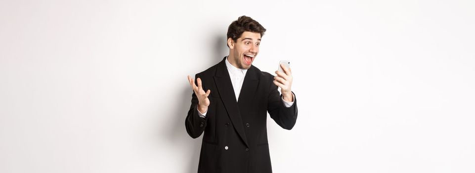 Portrait of happy smiling man in black suit, looking at smartphone screen with relieved and cheerful face, standing against white background.