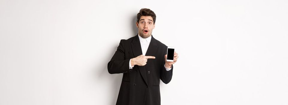 Portrait of attractive businessman in black suit, looking surprised and pointing finger at smartphone sreen, standing over white background.