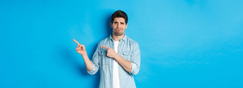 Portrait of skeptical adult guy pointing fingers right and smirking, exress disappointment and doubt, standing against blue background.