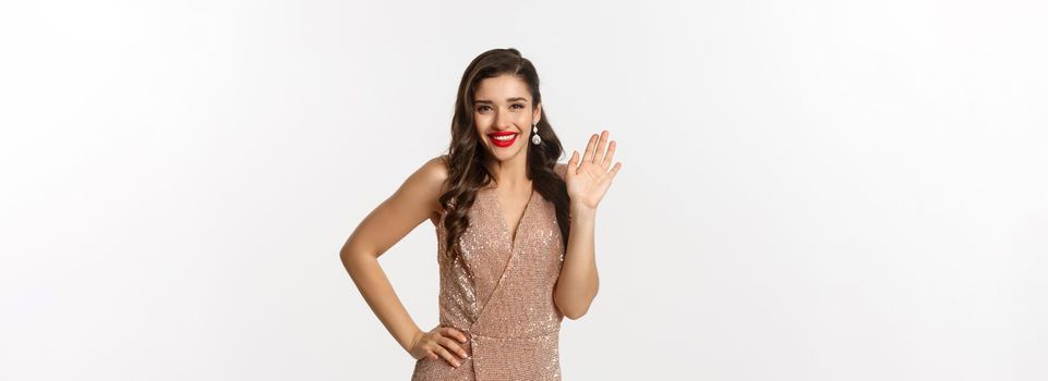 Christmas party and celebration concept. Happy beautiful woman in glamour dress saying hello, smiling and waiving hand with friendly expression, white background.