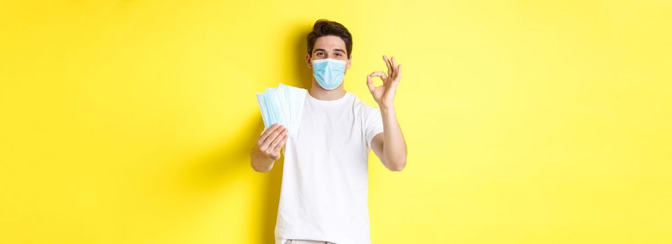 Concept of covid-19, quarantine and preventive measures. Satisfied man showing okay sign and giving medical masks, standing over yellow background.