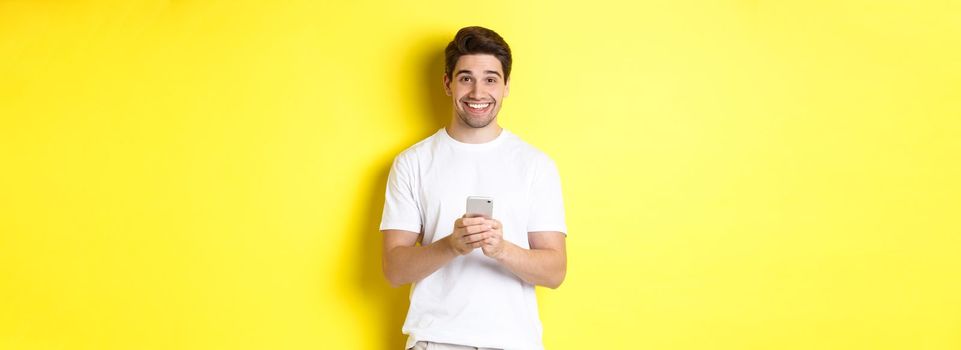 Man smiling and looking happy after reading promo offer on smartphone, standing against yellow background in white t-shirt.