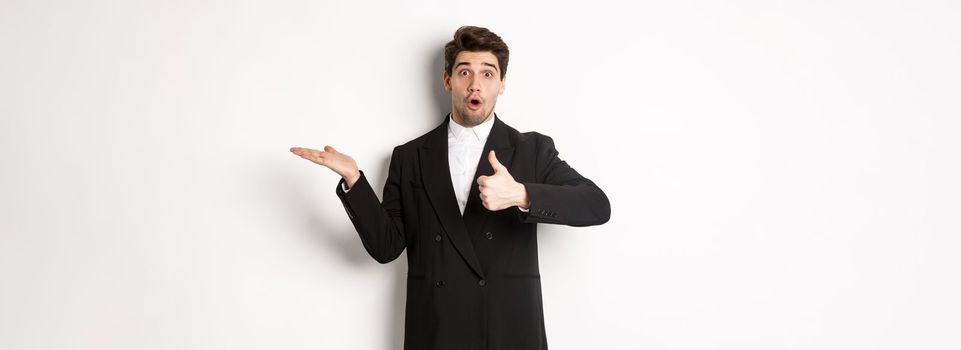 Portrait of hadnsome bearded man in formal suit, showing thumb-up and holding product in hand over white copy space, recommending product, standing over white background.