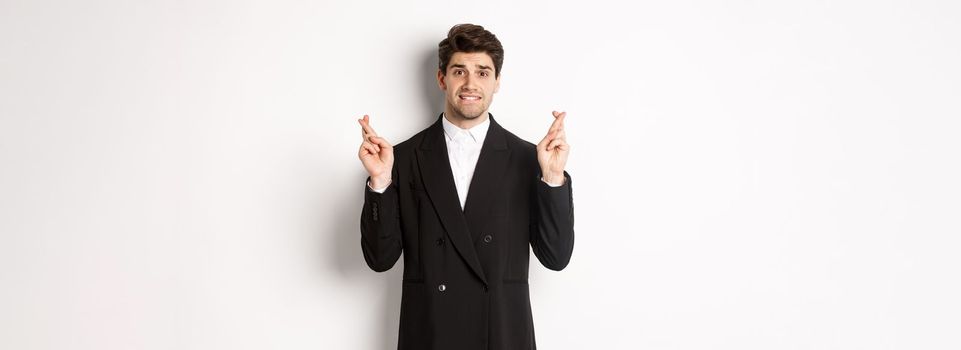 Nervous businessman in black suit crossing fingers, biting lip and making a wish, waiting for news, standing over white background hopeful.