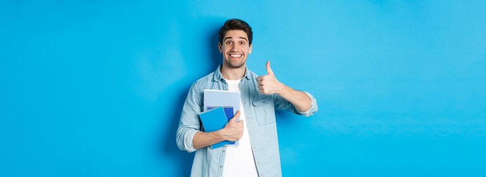 Happy young male model holding notebooks and showing thumb-up pleased, smiling and recommending courses, standing over blue background.