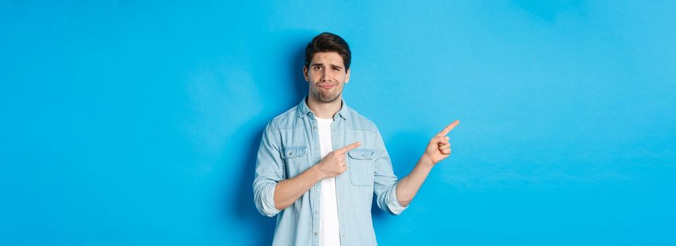 Skeptical adult man pointing fingers left, looking doubtful and unsure, standing over blue background.