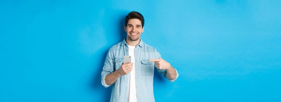 Concept of online shopping, applications and technology. Handsome man recommending app on smartphone, pointing at phone and smiling satisfied, standing over blue background.