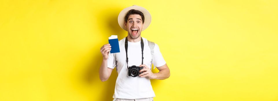 Travelling, vacation and tourism concept. Excoted tpirost showing passport with tickets, holding camera and wearing straw hat, standing over yellow background.