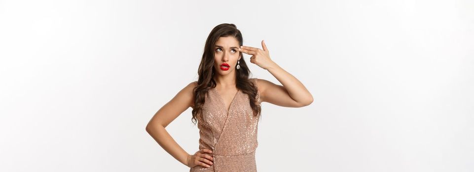 Celebration and party concept. Annoyed woman in elegant evening dress wants to kill herself from boredom, shooting finger gun in head and roll eyes irritated, standing over white background.