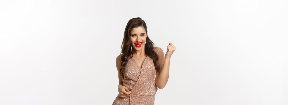 Celebration and party concept. Elegant young woman winning and feeling satisfied, wearing glamour dress, making fist pump and say yes, triumphing over white background.