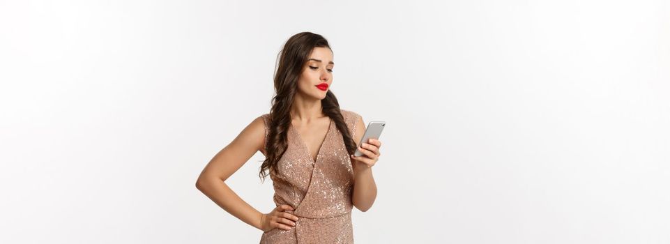 Christmas party and celebration concept. Careless woman in stylish dress reading message on mobile phone, looking skeptical, standing over white background.