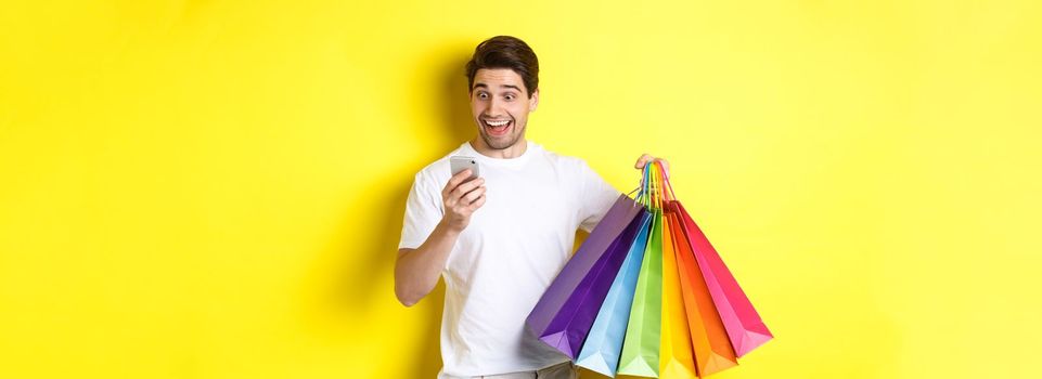 Excited man holding shopping bags and looking happy at mobile phone screen, standing over yellow background.