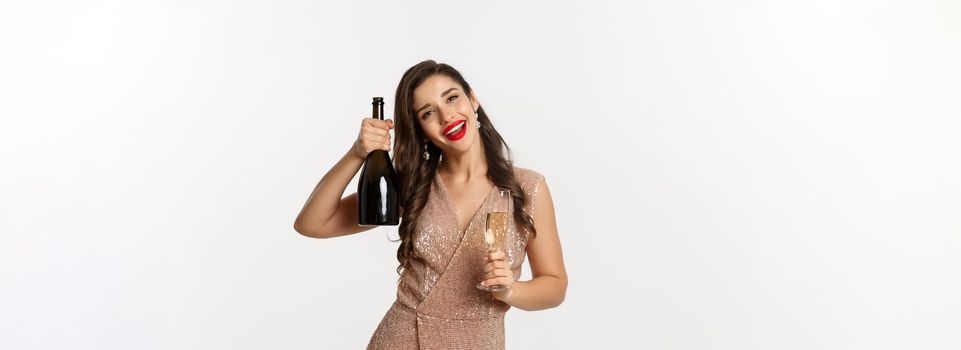 Winter holidays celebration concept. Happy woman on Christmas party saying cheers, raising bottle of champagne and smiling, standing in elegant dress, white background.